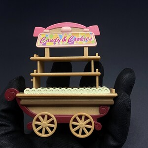 May include: A miniature candy and cookies cart with a pink and tan color scheme. The cart features two tan wheels, a red base, and a sign that reads "Candy & Cookies". The cart has shelves for displaying treats.
