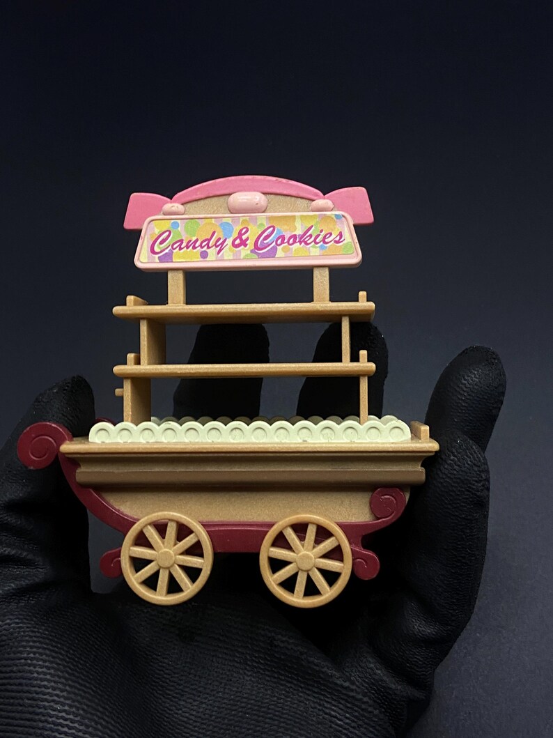 May include: A miniature candy and cookies cart with a pink and tan color scheme. The cart features two tan wheels, a red base, and a sign that reads "Candy & Cookies". The cart has shelves for displaying treats.
