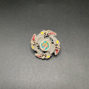 May include: A grey and red spinning top toy with a central emblem. The toy has a circular design with multiple blades and yellow and red accents. The centre has a circular emblem with a cartoon image.