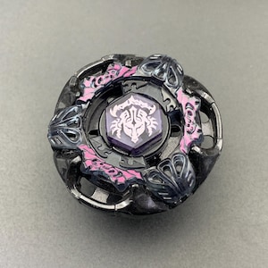 May include: A circular black and dark grey spinning top toy featuring a central hexagonal emblem with a white stylised graphic. The outer ring is accented with textured pink details and dark translucent plastic elements arranged in a symmetrical pattern.