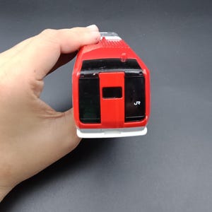 Może przedstawiać: Czerwony pociąg-zabawka z czarnymi oknami i białą podstawą. Pociąg ma z przodu wydrukowany numer "5". Pociąg-zabawka jest trzymany w dłoni osoby na czarnym tle.