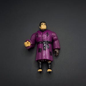 May include: A purple action figure of a man wearing a robe with a black belt. The figure has brown hair and a neutral expression. The robe has a textured pattern and patches. The figure's skin is tan, and the figure is standing.