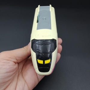 Puede incluir: Un tren de juguete con carrocería color crema, frontal negro y faros amarillos. La parte superior del tren es gris. El tren se sostiene en la mano de una persona sobre un fondo oscuro.