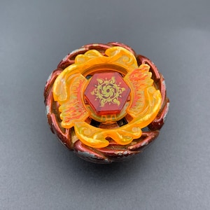 Puede incluir: Un juguete de peonza circular con un anillo exterior de color rojo oscuro metálico con desgaste visible. El centro consta de un componente naranja translúcido en forma de llama que rodea un perno hexagonal rojo con un emblema solar dorado.