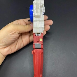 May include: A red, white, and blue toy with a handle. The toy has a blue wheel, a white body, and a red base. The toy is held in a hand against a dark background.