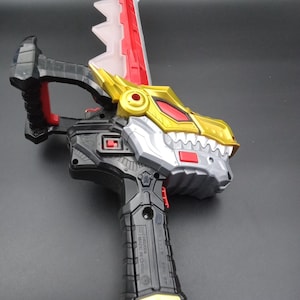 以下が含まれることがあります： 恐竜の頭のデザインの玩具の武器。おもちゃは主に黒、シルバー、ゴールドで、刃とハンドルに赤いアクセントがあります。ハンドルはテクスチャード加工されたグリップで、刃はギザギザのエッジです。