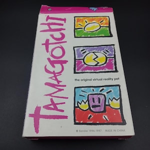 May include: A white rectangular box with the word "TAMAGOTCHI" in pink, stylized lettering. The box features three colorful illustrations and the text "the original virtual reality pet." The bottom of the box reads "© Bandai 1996-1997 MADE IN CHINA."