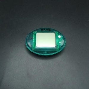 Op de afbeelding: Een turquoise elektronisch huisdierenspel met een rechthoekig scherm en knoppen. Japanse tekst is zichtbaar aan de bovenkant. Het scherm is leeg. Het apparaat is ovaal van vorm en heeft een transparante plastic behuizing.