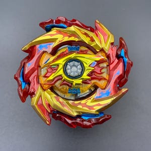 May include: A circular spinning top toy featuring a complex design with metallic gold, deep red, and bright blue accents. The centre has a silver circular component surrounded by layered, flame-like plastic blades in red and gold.