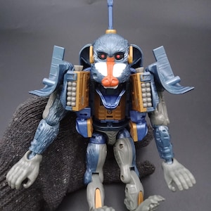 May include: A blue and gray action figure in the shape of a monkey with gold and orange accents. The toy has a long antenna and is posed with its arms and legs spread out. The figure is held by a gray glove.