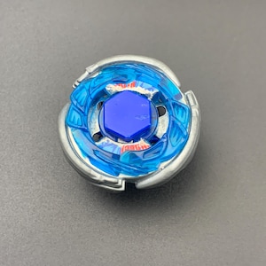 Puede incluir: Una peonza circular con un anillo exterior de metal plateado y una capa interior de plástico azul translúcido. El centro está dominado por un perno hexagonal de color azul oscuro, con pequeños detalles rojos visibles bajo el plástico.