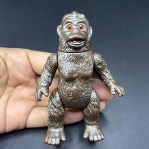 May include: A brown plastic toy gorilla with red eyes. The gorilla is in a standing position with its arms at its sides. The toy has a detailed texture and is likely a vintage collectible.