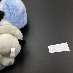 May include: A white and blue plush toy dog wearing a blue hat. The toy has black eyes and a black nose. The toy is sitting on a black surface. The toy has a tag that says "PEANUTS C 2001 United Features Syndicate, Inc. Made in China."