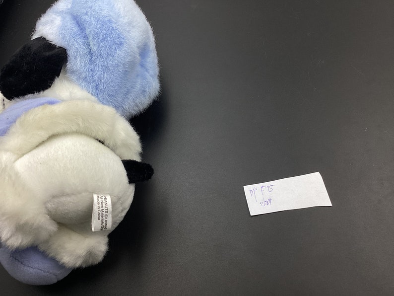 May include: A white and blue plush toy dog wearing a blue hat. The toy has black eyes and a black nose. The toy is sitting on a black surface. The toy has a tag that says "PEANUTS C 2001 United Features Syndicate, Inc. Made in China."