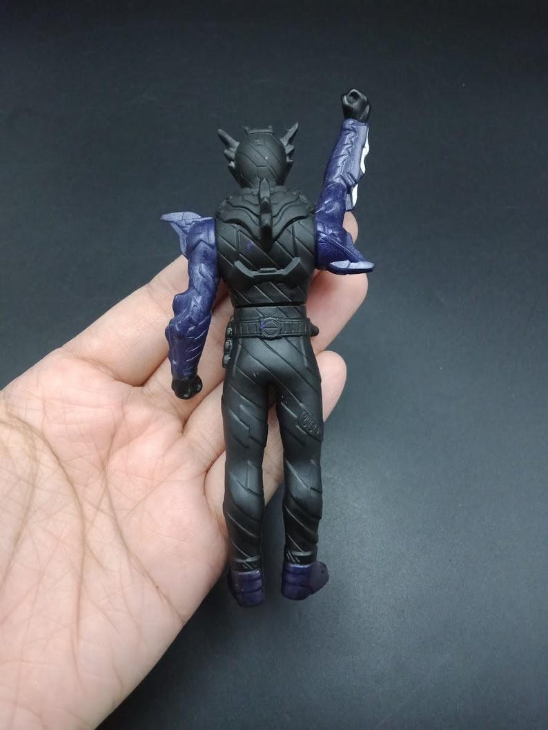 May include: A black action figure with purple accents, viewed from the back. The figure has a detailed design, including a belt, textured legs, and a raised arm. The figure's arms and shoulder blades are purple.