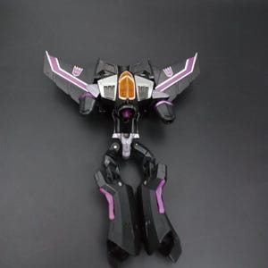 May include: A black and purple toy robot in a jet fighter configuration. The toy has purple accents on the wings and legs, with a silver and orange cockpit. The wings have a white stripe and a purple emblem.