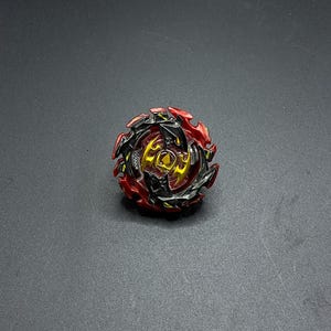 K&ouml;nnte beinhalten: Ein roter, schwarzer und goldener Kreisel mit einem scharfen, kantigen Design. Der Kreisel hat einen zentralen Kern mit einem kreisf&ouml;rmigen Design und einen Zahnkranz am Rand.