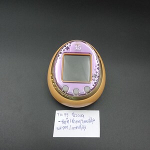 May include: A vintage, egg-shaped digital pet device with a purple screen and gold trim. The device has a small screen and three buttons. The top of the device has the number "15" and star-shaped designs. A handwritten note is below the device.