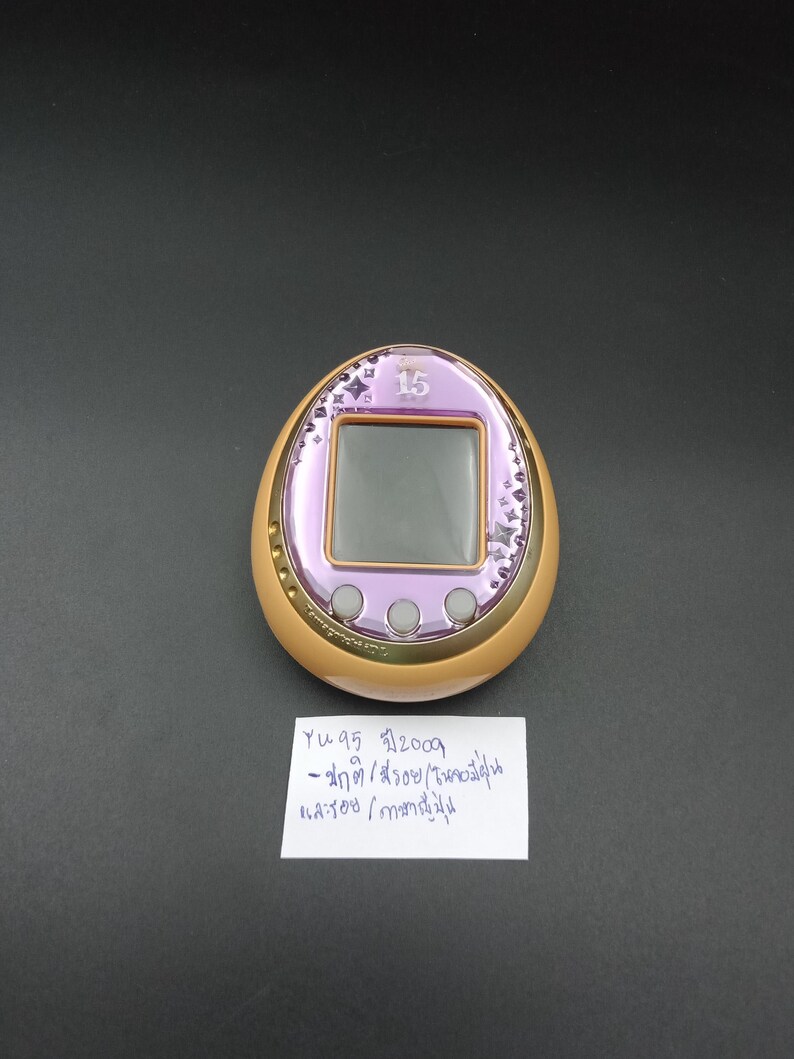 May include: A vintage, egg-shaped digital pet device with a purple screen and gold trim. The device has a small screen and three buttons. The top of the device has the number "15" and star-shaped designs. A handwritten note is below the device.