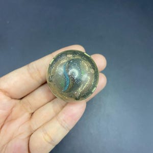 Könnte beinhalten: Eine klare, runde Murmel mit einem wirbelnden blau-goldenen Design im Inneren. Die Murmel hat kleine, gleichmäßig verteilte Löcher am Rand. Die Murmel wird in einer Hand vor einem dunklen Hintergrund gehalten.