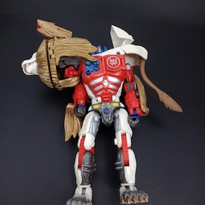 May include: A red, white, and blue action figure with a lion-like head and fur details. The figure has articulated joints and a detailed design, with a symbol on its chest. The toy is standing on a dark surface.