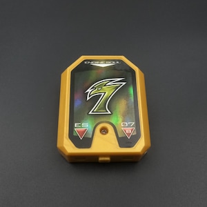 Può includere: Un giocattolo Engine Soul in plastica gialla con un pannello olografico centrale che presenta un logo verde con il numero sette. Il dispositivo mostra il testo ENGINE SOUL in alto, con le etichette ES e 07 B ai lati di una piccola luce.
