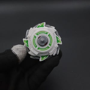 May include: A white and grey toy with green detailing and the word "DRAGOONG" printed on it. The toy has a circular design with a central grey piece and green triangular shapes around the edge. The toy is held by a person wearing a black glove.