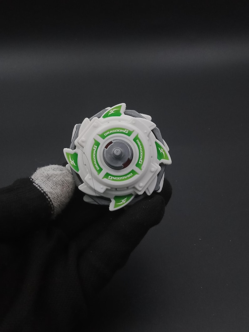 May include: A white and grey toy with green detailing and the word "DRAGOONG" printed on it. The toy has a circular design with a central grey piece and green triangular shapes around the edge. The toy is held by a person wearing a black glove.