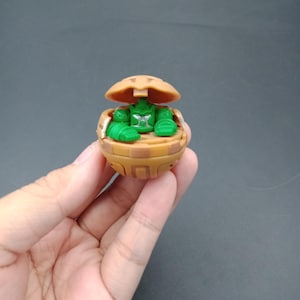May include: A brown and green toy figure, held in a hand, is open to reveal a green character inside. The toy is spherical with a camouflage pattern and a hinged top. The character has green arms and legs.
