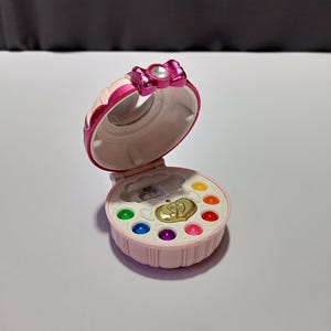May include: A round, light pink plastic compact case with a hinged lid. The lid features a metallic pink bow with a central clear gem. The interior contains a gold heart-shaped emblem and six small, colourful circular beads in green, blue, purple, pink, orange, and yellow.