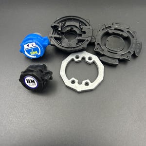 May include: A disassembled toy, featuring various components in black, blue, and silver. The parts include a blue piece with white text, two black circular pieces, a silver ring, and a black piece with white text. The toy is designed for play.