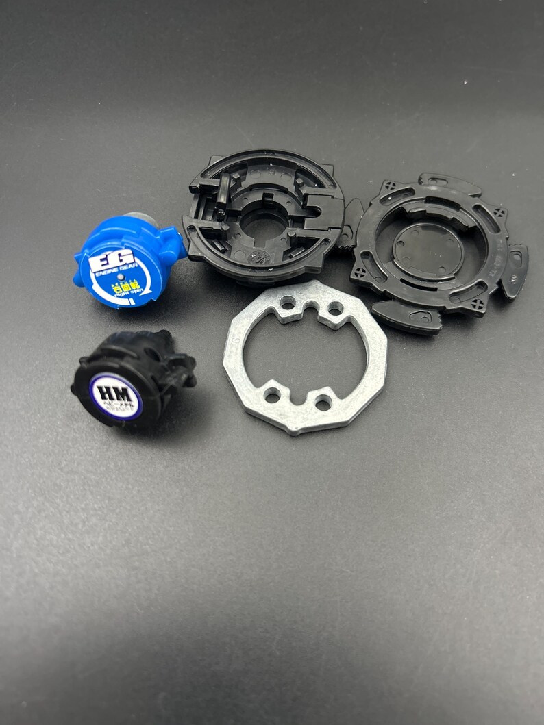 May include: A disassembled toy, featuring various components in black, blue, and silver. The parts include a blue piece with white text, two black circular pieces, a silver ring, and a black piece with white text. The toy is designed for play.