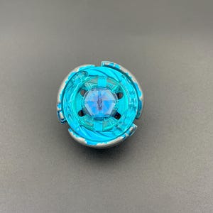 Puede incluir: Un juguete de peonza circular con un centro de plástico translúcido de color cian brillante y un emblema hexagonal. El anillo exterior está construido con componentes de plástico plateado metálico y cian, dispuestos en un diseño asimétrico de tres puntas.