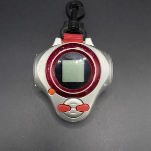 Peut inclure: Un appareil numérique portable argenté avec des détails rouges et noirs. Il comporte un petit écran, deux boutons rouges et un bouton orange. Un clip noir est attaché en haut avec une sangle rouge.