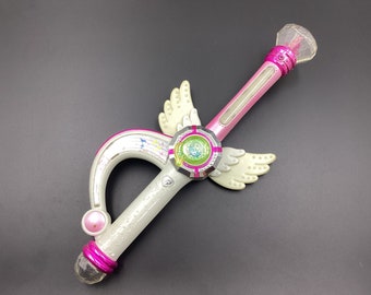 May include: A pink and white toy wand with a clear, faceted tip. The wand features a curved handle, wings, and a circular design with a green center. The wand is approximately 12 inches long.