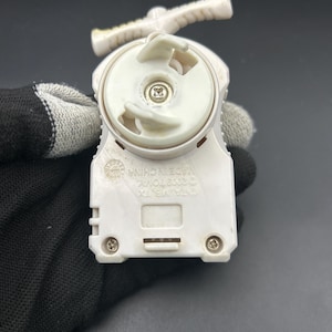 Puede incluir: Un juguete de plástico blanco y beige con un asa y una parte superior circular. El juguete tiene una pequeña ranura rectangular y está sujeto por una mano enguantada. El juguete tiene el texto "©OTA,VB,TX MADE IN CHINA ©2002 TOMY" impreso.