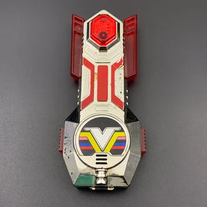 Op de afbeelding: Een zilverkleurig, zwart en rood plastic speelgoedvoertuig met een opvallend V-logo met horizontale strepen in geel, roze, blauw en rood. Het speelgoed heeft doorschijnende rode zijpanelen en een zeshoekig rood gedeelte aan de bovenzijde.