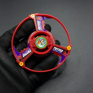 May include: Red and purple plastic Beyblade with a scorpion design and the text "TRYPIO BEYBLADE FLYING" on the side.