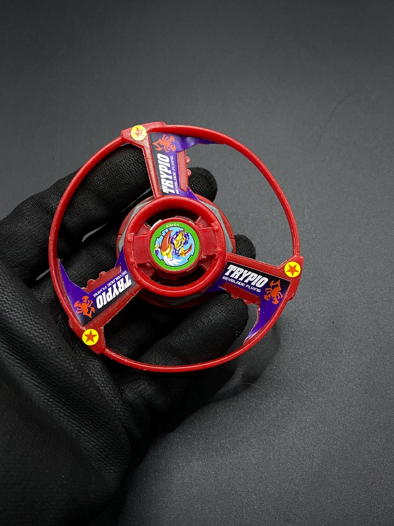 May include: Red and purple plastic Beyblade with a scorpion design and the text "TRYPIO BEYBLADE FLYING" on the side.
