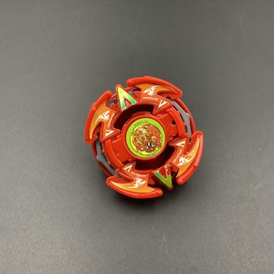 May include: A red and orange spinning top toy featuring a circular central emblem with a detailed graphic illustration, surrounded by four symmetrical wing-like protrusions with green accents and small white dragon-shaped logos printed on the surface.