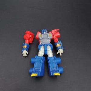 May include: A blue, red, and gray toy robot figure. The toy has blue legs, a gray torso, red arms, and a blue head. The robot has yellow and white accents. The toy is a Transformer.