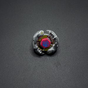 Może przedstawiać: Srebrny i czerwony Beyblade z niebieskim i żółtym wzorem na centralnym elemencie. Beyblade znajduje się na czarnym tle.