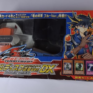 Puede incluir: Un set de Yu-Gi-Oh! 5D's Duel Disk DX en caja. La caja es roja, azul y naranja, con una ilustración de personaje y cuatro cartas coleccionables. El set incluye un Duel Disk y un protector de cartas.