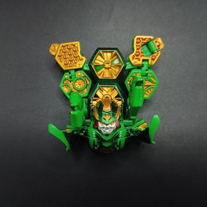 May include: Green and gold toy robot with articulated limbs and a detailed face. The toy has a complex design with hexagonal and geometric shapes. The robot's head features red and white accents, and the overall aesthetic is futuristic.