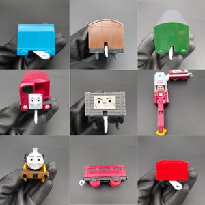 May include: A selection of toy train carriages in different colours and styles. The set features a blue boxcar, a brown covered wagon, a green hopper car, a red engine, a grey flatbed car with a face, a red and white crane car, a yellow and brown engine, a red flatbed car, and a red boxcar.