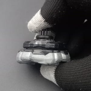 Pu&ograve; includere: Una trottola giocattolo nera e grigia con un design strutturato. La parte superiore ha una struttura a forma di ingranaggio nella parte superiore e una base grigia a forma di pietra. Il giocattolo &egrave; tenuto da un guanto nero e grigio.