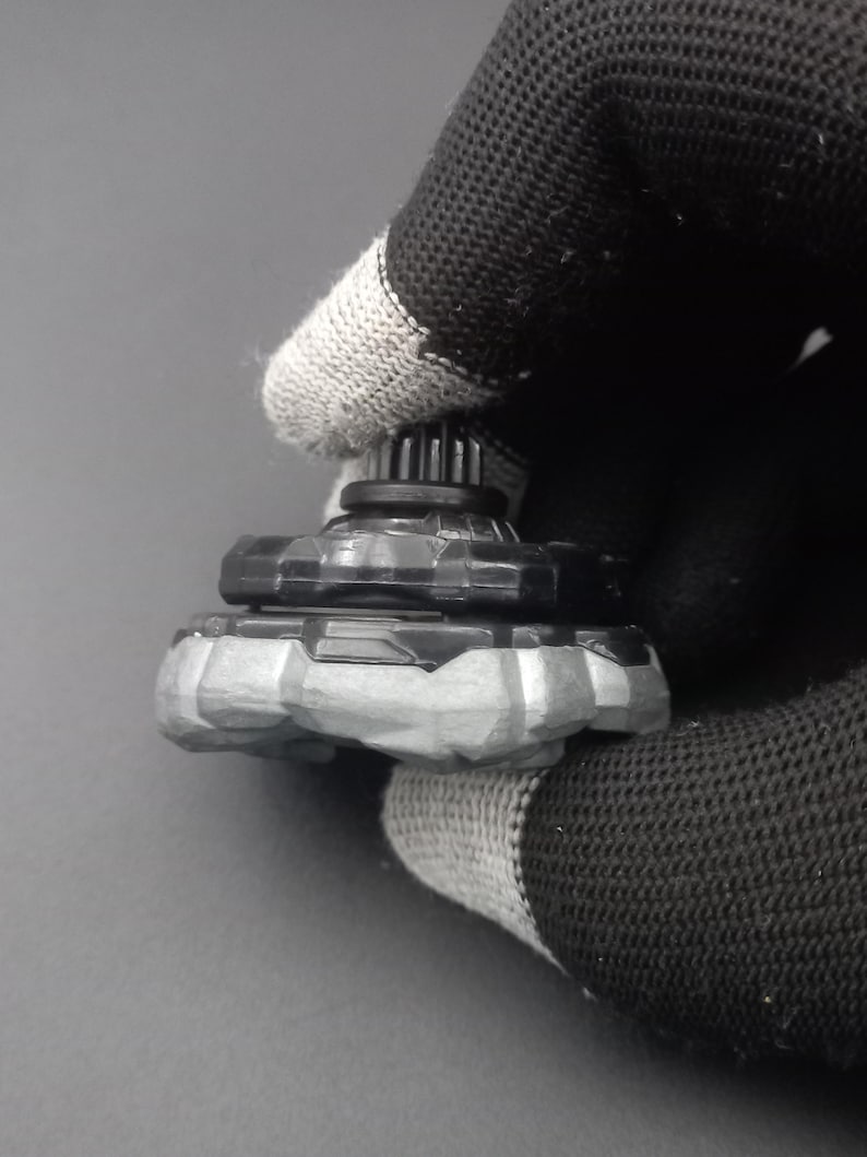 Pu&ograve; includere: Una trottola giocattolo nera e grigia con un design strutturato. La parte superiore ha una struttura a forma di ingranaggio nella parte superiore e una base grigia a forma di pietra. Il giocattolo &egrave; tenuto da un guanto nero e grigio.