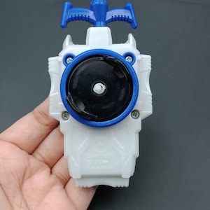 Puede incluir: Un juguete blanco y azul con un centro negro y un asa azul. El juguete tiene un diseño circular con un anillo azul alrededor del centro negro. El asa es azul y tiene una forma única. El juguete se sostiene en una mano.