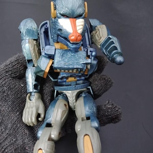 以下が含まれることがあります： オレンジとゴールドのアクセントが付いた、青とグレーのロボットマンドリルのアクションフィギュア。 おもちゃは可動式の関節と詳細なデザインで、毛皮のような質感があります。 フィギュアは灰色の手袋で保持されています。