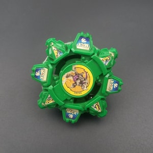 May include: A green and blue toy with a circular design. The toy has a central image of a character and the word "MAX" on several sections. The toy is on a gray surface.
